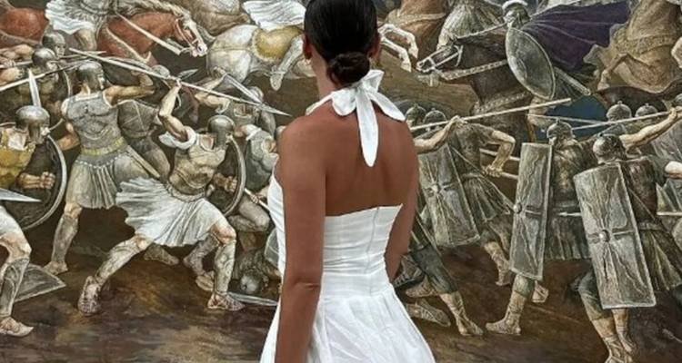 Personne en robe blanche debout devant une fresque représentant une scène de bataille.
