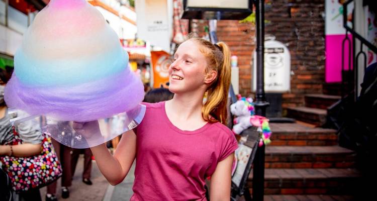 Girl holding a large rainbow-colored cotton candy in a bustling street.