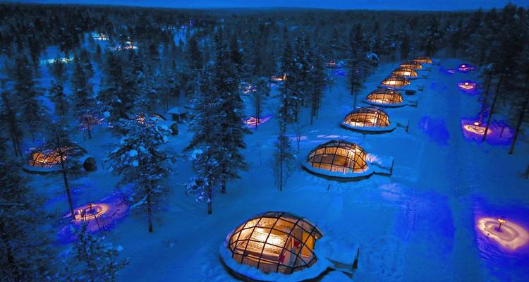 Aerial view of illuminated glass igloos in a snowy forest at night.