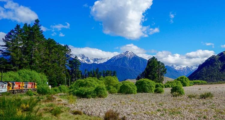 Un train de voyageurs serpente à travers une forêt luxuriante avec des pics enneigés sous un ciel bleu éclatant