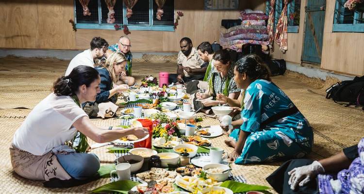 Reizigers zitten met gekruiste benen op geweven matten en delen een traditioneel gemeenschappelijk diner in een dorpshuis.