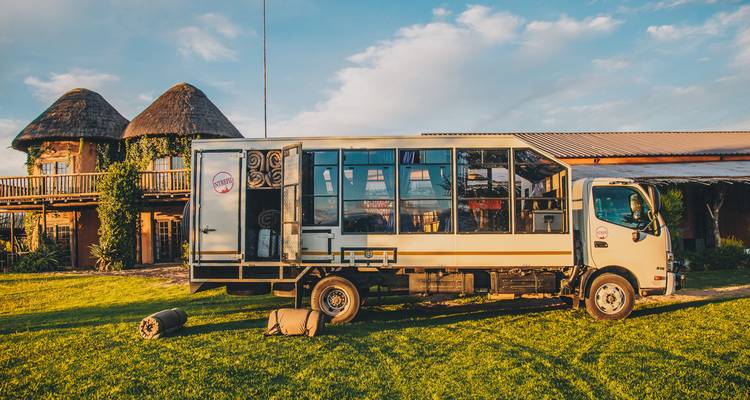 An overland truck parked on a grassy area with tents nearby and a building in the background.