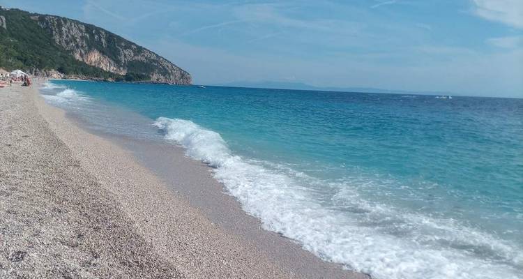 Plage de galets avec mer turquoise et montagnes en arrière-plan.