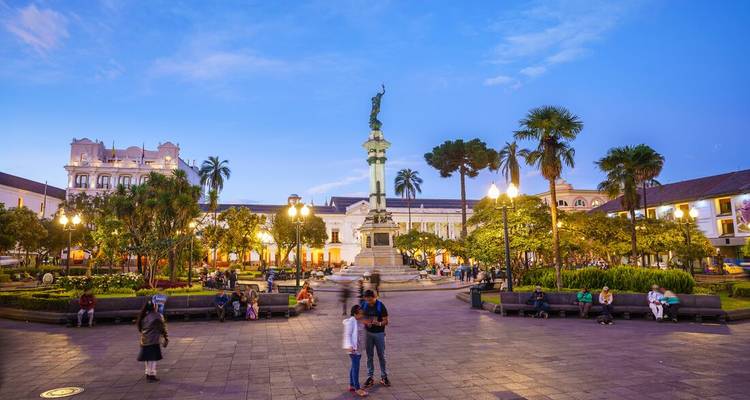Een plein met een standbeeld, omringd door mensen en weelderig groen in de avond.