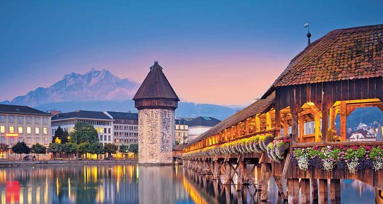 A charming wooden bridge with flowers, a tower, and mountains in the background reflecting in a river.