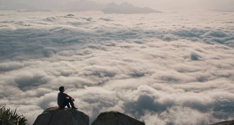 Persoon zittend op een rots boven een zee van wolken.