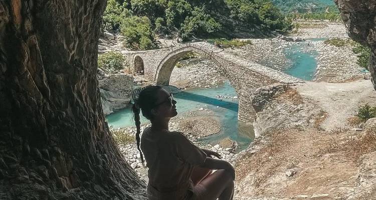 Femme assise dans une grotte surplombant un pont de pierre historique.