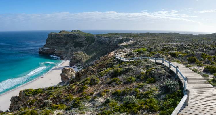 Houten loopbrug door kustvegetatie naar een strand.