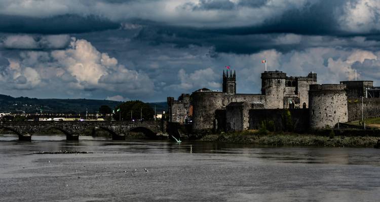 Un château de pierre historique au bord d'une rivière sous un ciel nuageux.