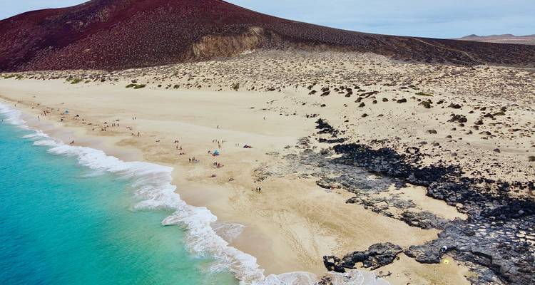 Vue aérienne d'une plage de sable, d'eaux claires et de personnes profitant du soleil.