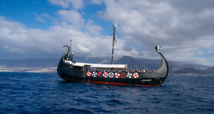 Bateau de style viking naviguant sur la mer avec des montagnes en arrière-plan.