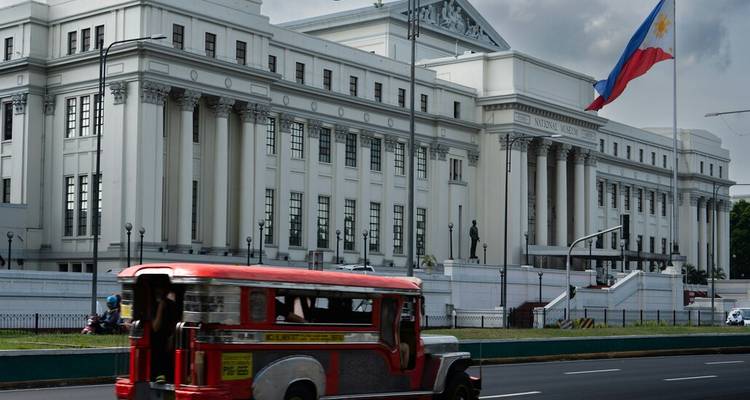 Un jeepney rouge classique passe devant la grande façade blanche du Musée national des Philippines avec le drapeau national qui flotte.