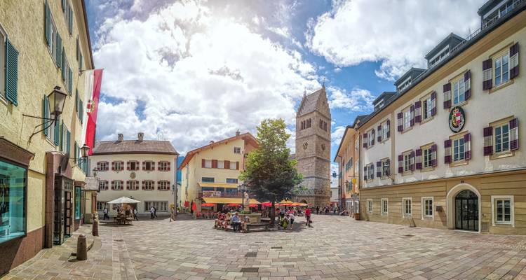 Una plaza encantadora con edificios, una torre del reloj y una bandera austriaca, donde se ve gente relajándose.