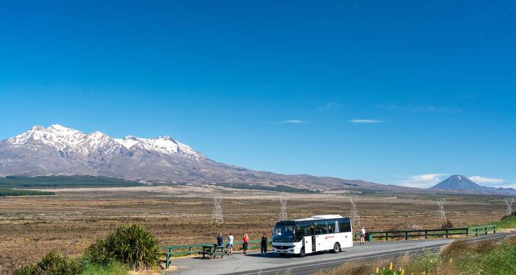 Reisebus geparkt an einem Aussichtspunkt am Straßenrand mit schneebedeckten Gipfeln des Tongariro-Nationalparks, die sich hinter weiten Ebenen erheben.