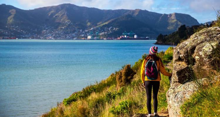 Rückansicht eines Wanderers mit bunter Mütze, der entlang eines Küstenpfads geht und auf eine weite blaue Bucht und eine entfernte Stadt blickt.