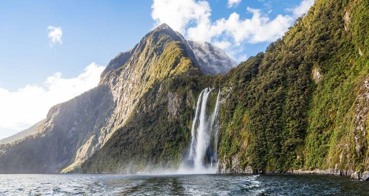 Dramatische steile Klippen und ein hoher Wasserfall, der in die Fjordgewässer von Milford Sound unter hellem Himmel stürzt.