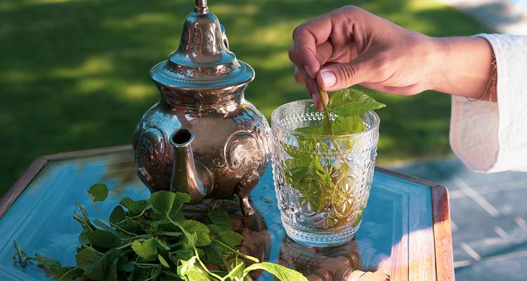 Hand pouring mint tea from a silver teapot into a glass.