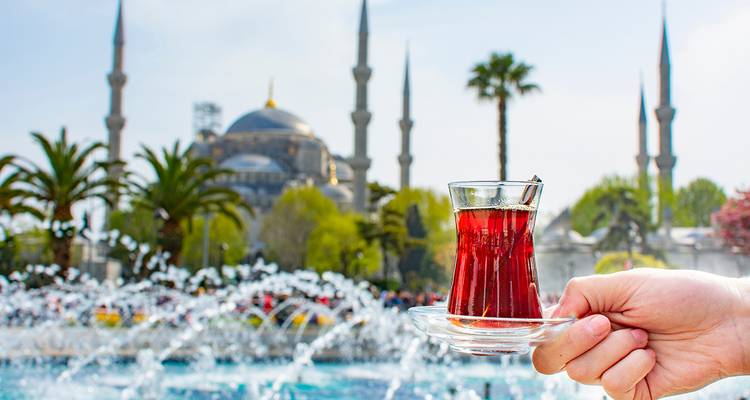Une personne tenant un verre de thé avec une grande mosquée et une fontaine en arrière-plan.