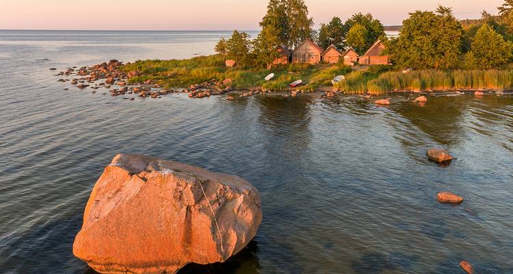 Un rivage rocheux avec un rocher géant dans l'eau au coucher du soleil, avec des maisons rustiques et de la verdure.