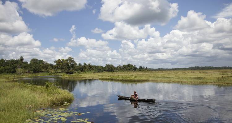 Person in a canoe on a tranquil river surrounded by lush landscape under a blue sky with clouds.
