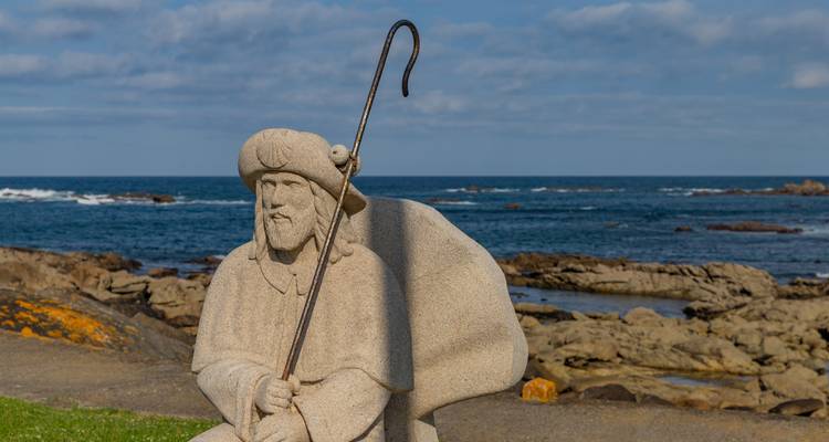 Statue de pierre d'un pèlerin surplombant l'océan avec une côte rocheuse.