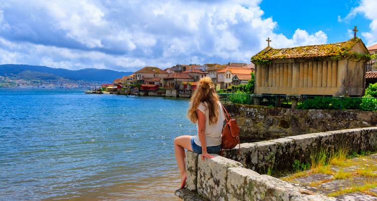 Femme assise sur un mur de pierre au bord de l'eau, surplombant une ville historique.