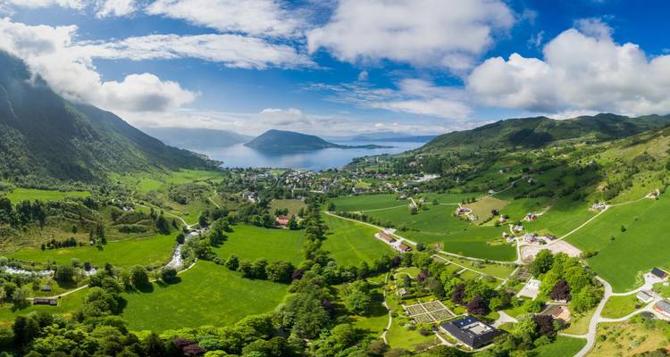 Vista aérea soleada de un pintoresco pueblito de fiordo rodeado de verdes colinas ondulantes.