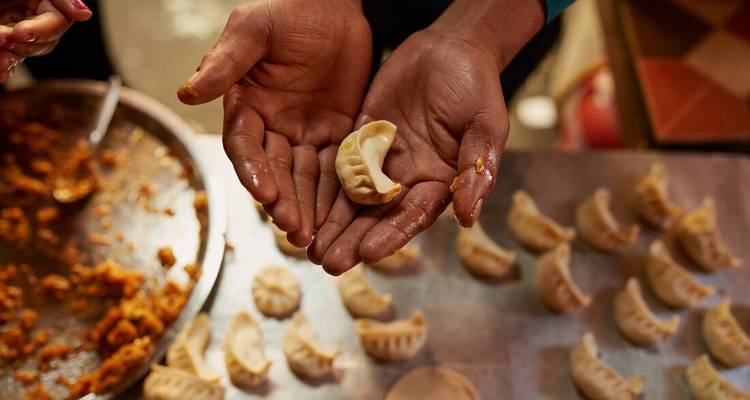 Des mains façonnant des boulettes fraîches sur une table en métal entourée de rangées de momos non cuits.