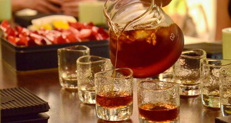 A hand pouring coffee into small glasses with a fruit platter in the background.