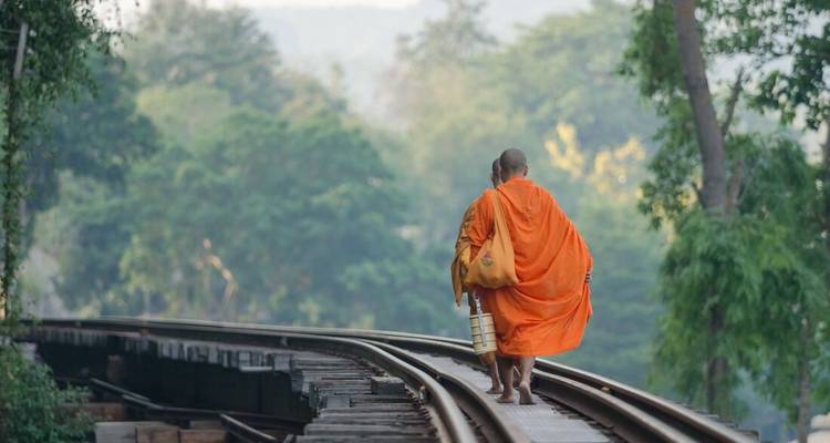 Two monks walking along railway tracks surrounded by lush forest.