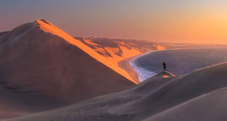 A lone traveller stands atop glowing orange sand dunes that plunge dramatically into the Atlantic Ocean at sunset.
