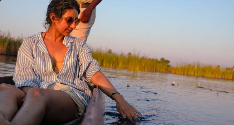 Woman relaxes in a traditional mokoro canoe, trailing her hand in the calm Okavango Delta waters at dusk.