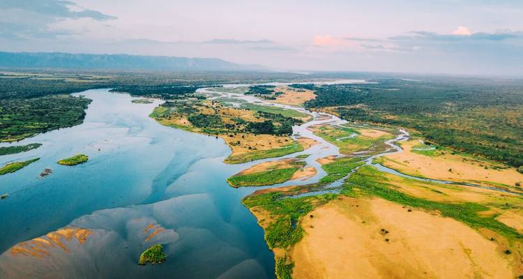 Drone view of winding channels and islands of the Okavango Delta meeting a wide river under pastel skies.