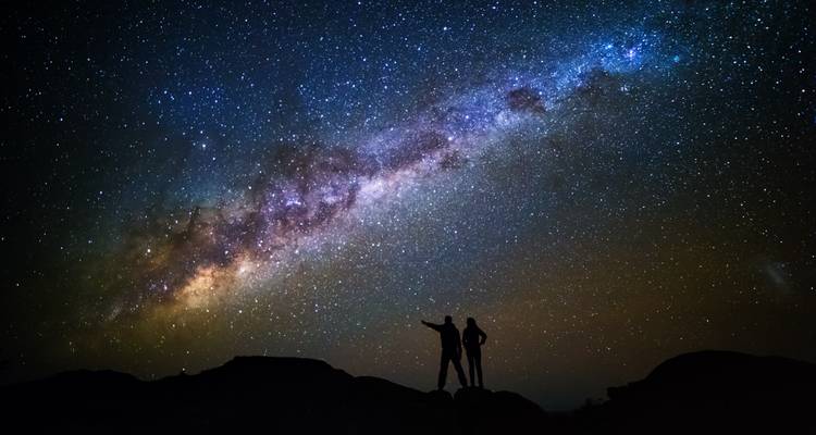 Silhouetted travellers point toward the luminous Milky Way stretching across a dark desert sky.