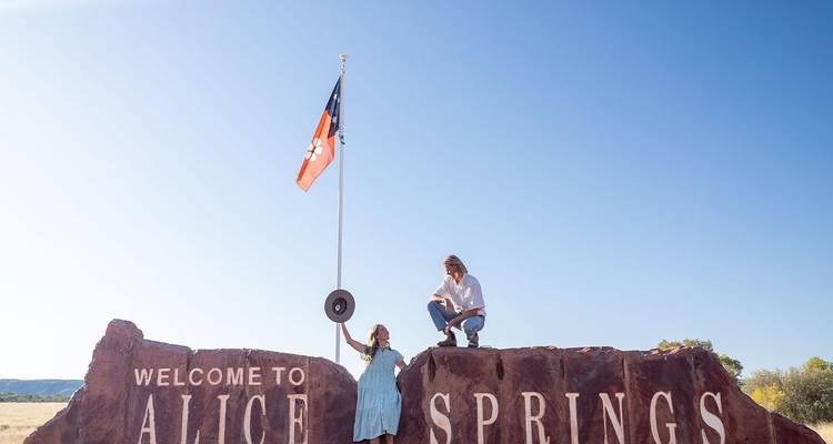 Dos viajeros posando en la gran roca roja "Bienvenido a Alice Springs" bajo un cielo azul despejado