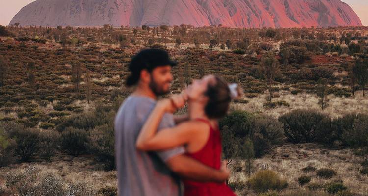 Un couple s'enlace au premier plan avec Uluru qui brille en rouge au coucher du soleil en arrière-plan.