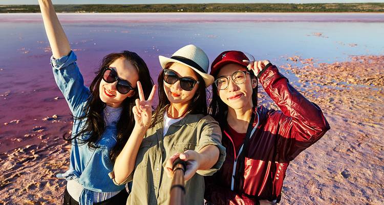Trois jeunes femmes prennent un selfie joyeux avec un lac rose vif et un horizon plat qui s'étendent derrière elles.
