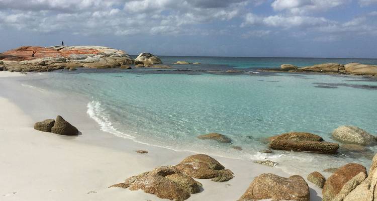 Plage de sable blanc immaculé avec des eaux turquoise peu profondes et des rochers distinctifs couverts de lichen orange sous un ciel partiellement nuageux.