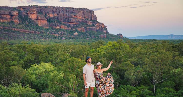 Couple stands on a rocky lookout above vast green woodland with towering red sandstone escarpment at dusk in the outback.