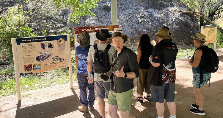 Small tour group reads interpretive signs on a shaded walkway in a national park setting.