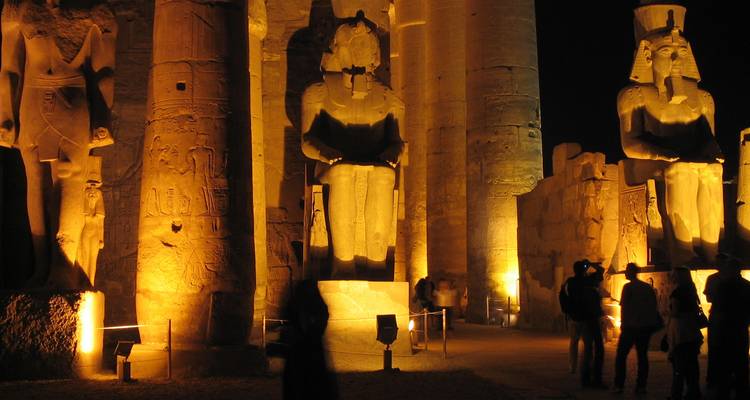Estatuas faraónicas colosales del Templo de Luxor iluminadas por luces doradas en la noche con visitantes abajo.