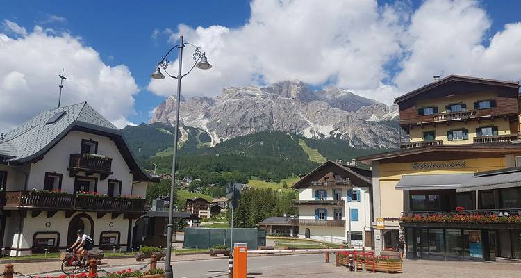 Plaza de pueblo alpino con ciclistas, chalets y el dramático telón de fondo de las montañas Dolomitas.
