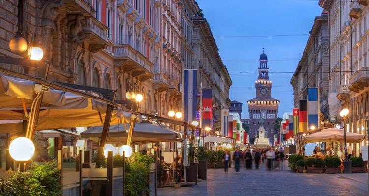 Calle peatonal nocturna en Milán llena de cafés que lleva al Castillo Sforza.