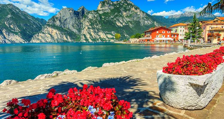 Paseo bordeado de flores junto al Lago de Garda con acantilados imponentes y casitas encantadoras de pueblos junto al lago.