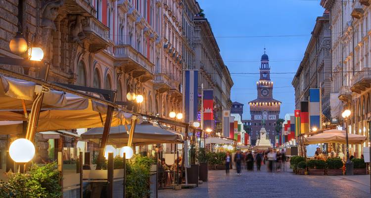 Une rue de ville animée avec une vue sur une tour au loin, probablement à Milan.