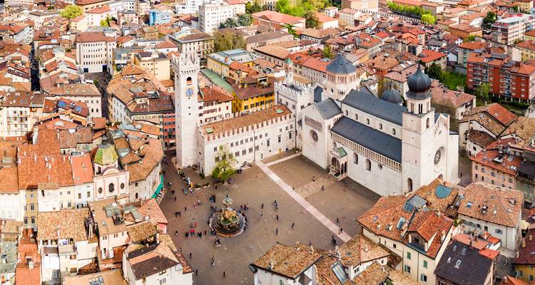 Aerial panorama of an Italian town square with a grand cathedral, clock tower, surrounding terracotta-roofed buildings and a central fountain.