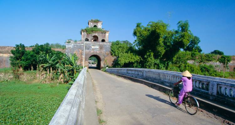 A person cycling over a bridge towards an ancient gate.