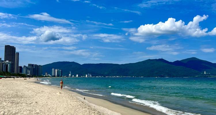 Sandy beach with promenading person, ocean waves, and city skyline against mountains.