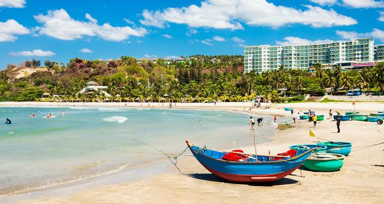 Escena de la playa soleada de Mui Ne con coloridas barcas de canasta redondas y un barco de pesca de madera azul en aguas turquesas respaldadas por resorts rodeados de palmeras.