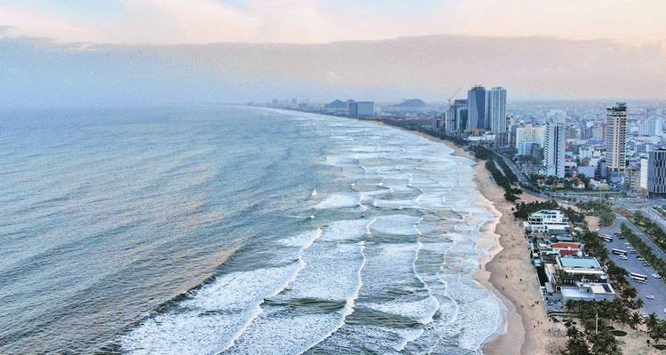 Vista aérea de la larga costa arenosa de Da Nang y el horizonte de la ciudad al amanecer, con olas rodando hacia la orilla.